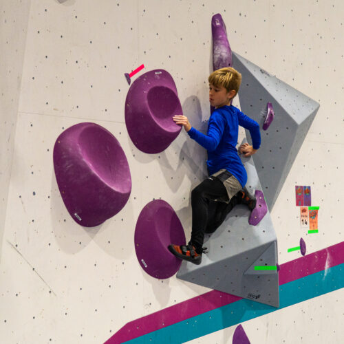 Young boy climbing an indoor bouldering wall using large purple holds and a grey volume, focusing on balance at Flashpoint Easton