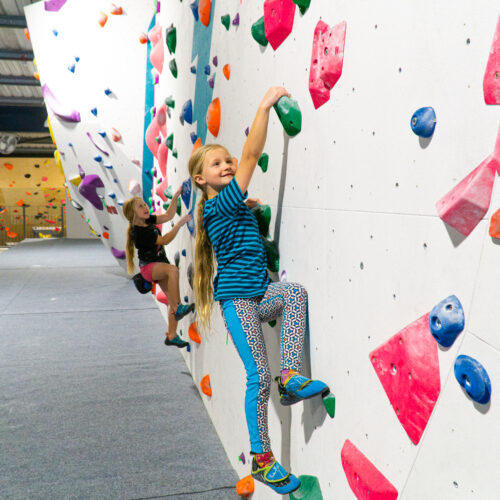 Two girls climbing an indoor bouldering wall with green, blue and pink holds, smiling as they move along the wall at Flashpoint Easton