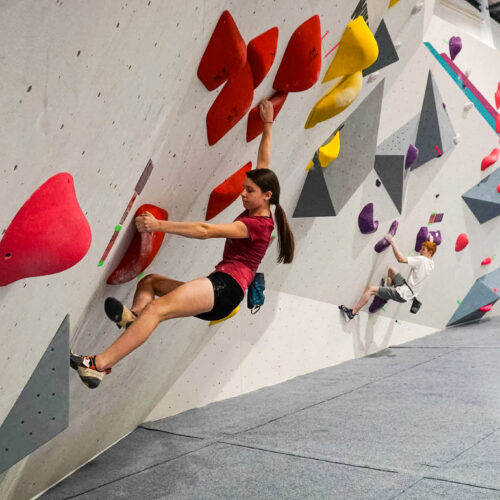 Two climbers on an indoor overhanging bouldering wall using red holds, moving across the wall on separate routes at Flashpoint Easton