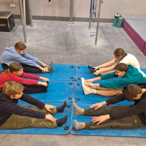 Group of young climbers sitting on mats and stretching their legs in a circle during a warm-up session at Flashpoint Easton