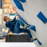Boy climbing low on a bouldering wall using large blue volumes and an orange hold, wearing climbing shoes at Flashpoint Easton
