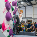 Man climbing on a bouldering wall using large purple volumes and green holds while a crowd watches behind at Flashpoint Easton