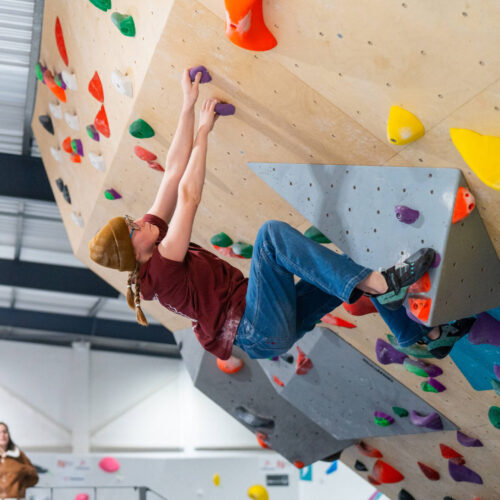 Woman climbing on a steep overhang, reaching for purple holds while balancing on a grey volume at Flashpoint Easton