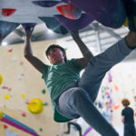 Man hanging from large holds on an overhanging indoor climbing wall with legs extended while others watch below at Flashpoint Easton