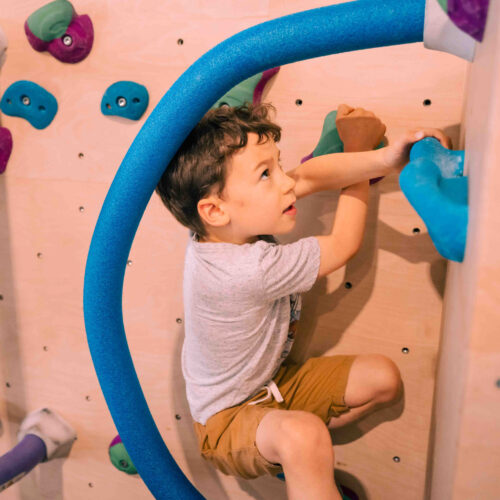 Boy bouldering on the kids’ wall while reaching for a blue hold inside a foam hoop at Flashpoint Easton