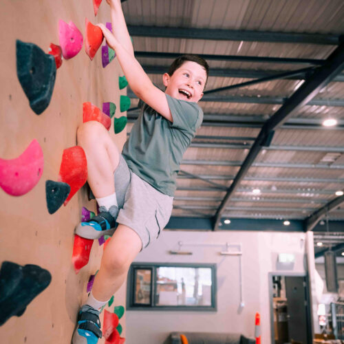 Boy climbing high on the kids’ wall using red and pink holds while laughing at Flashpoint Easton