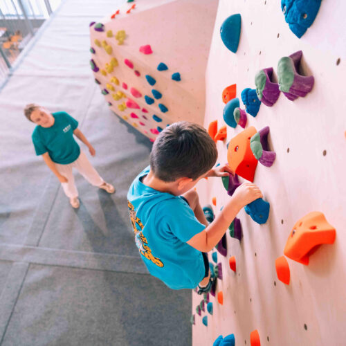 Boy climbing on colourful holds on a kids’ wall while a Flashpoint Easton staff member looks on from the mats