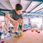 Boy stepping onto yellow holds at the top of a low kids’ climbing wall while adults watch in the background at Flashpoint Easton